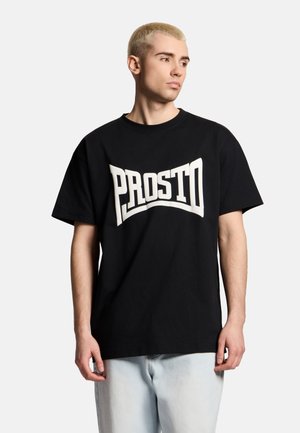 Młody mężczyzna z krótkimi blond włosami, w czarnej oversize koszulce z napisem "PROSTO" dużymi białymi literami, w połączeniu z jasnoniebieskimi dżinsami.