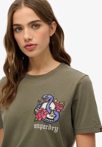 Olivengrønt T-shirt med et fremtrædende broderet design af en blå slange, der snor sig omkring et kranium og lyserøde roser. Teksten "superdry" nederst.