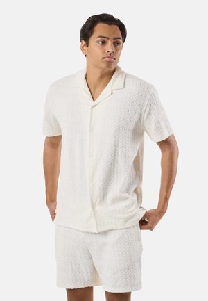 Jonge man die een witte, gestructureerde, korte mouwen blouse met knopen en bijpassende korte broek draagt, staat met zijn handen in de zakken en kijkt opzij.