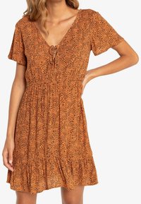 Robe orange à motif léopard, manches courtes, encolure en V avec un détail noué, et ourlet à volants à plusieurs niveaux. Fabriquée en tissu léger.