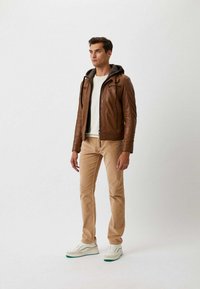 Bruine leren jas met een grijze capuchon, een crèmekleurig shirt eronder, beige broek en witte sneakers met groene accenten, staande tegen een effen achtergrond.