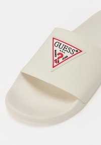 Sabot alb cu un suport texturat pentru picior și o curea lată cu o patch cu logo triunghiular "GUESS" în culorile roșu și negru.