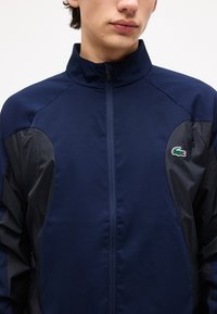 Νέος άνθρωπος που φοράει ένα μπλε σκούρο μπουφάν με φερμουάρ, με μαύρα πάνελ και μικρό πράσινο λογότυπο κροκοδείλου στο στήθος.