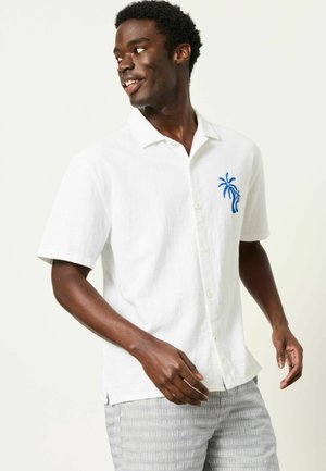 Lächelnder Mann trägt weißes Kurzarm-Hemd mit blauem Palmen-Stick, schaut nach links, kombiniert mit hellgrauen gestreiften Hosen.