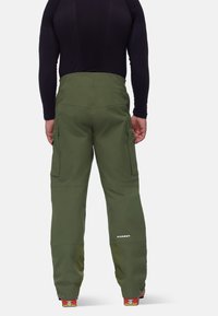 Pantalons de ski imperméables verts avec poches zippées, tissu renforcé en bas et sections en maille de ventilation, associés à un haut noir à manches longues.