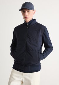 Navy padded jacket with a high collar, front zip closure, side pockets, and ribbed cuffs, paired with a matching cap.