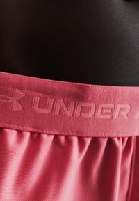 Κοντινό πλάνο ροζ μέσης Under Armour σε αντίστοιχα ροζ αθλητικά σορτς με σκοτεινό φόντο.
