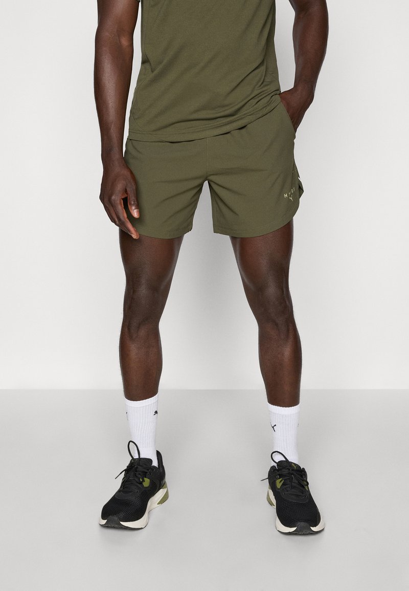 Shorts de sport en vert olive avec poches latérales, associés à des chaussures de sport noires et des chaussettes blanches à la cheville. Tissu lisse, coupe décontractée.