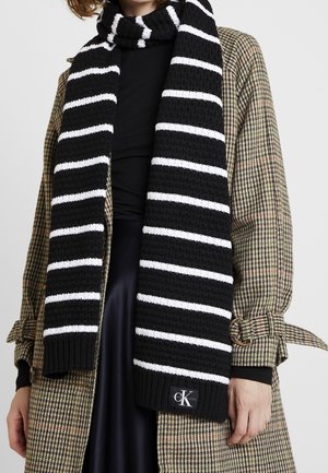 Écharpe en maille rayée noire et blanche avec logo Calvin Klein portée sur un manteau à carreaux ceinturé et un haut noir.