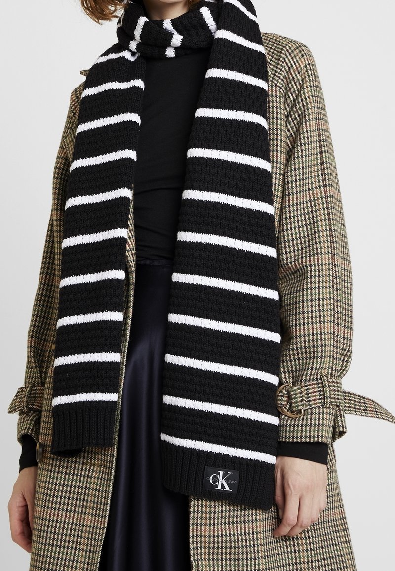 Écharpe en maille rayée noire et blanche avec logo Calvin Klein portée sur un manteau à carreaux ceinturé et un haut noir.