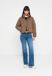 Femme se tenant contre un fond blanc, portant une doudoune marron, un haut noir, un jean flare bleu et des baskets blanches, les mains dans les poches.