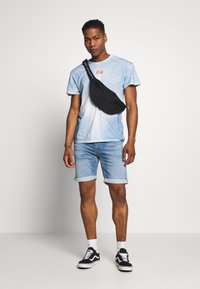 Modrá tie-dye trička s krátkým rukávem, světle modré džínové šortky, černý ledvinka s popruhem s logem, bílo-černé tenisky, ohrnuté šortky.