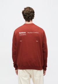 Άντρας με σγουρά μαλλιά στέκετ�αι με την πλάτη γυρισμένη, φορώντας μία καφέ χνουδωτή μπλούζα με λευκά γράμματα στην πλάτη και ανοιχτό μπεζ παντελόνι.