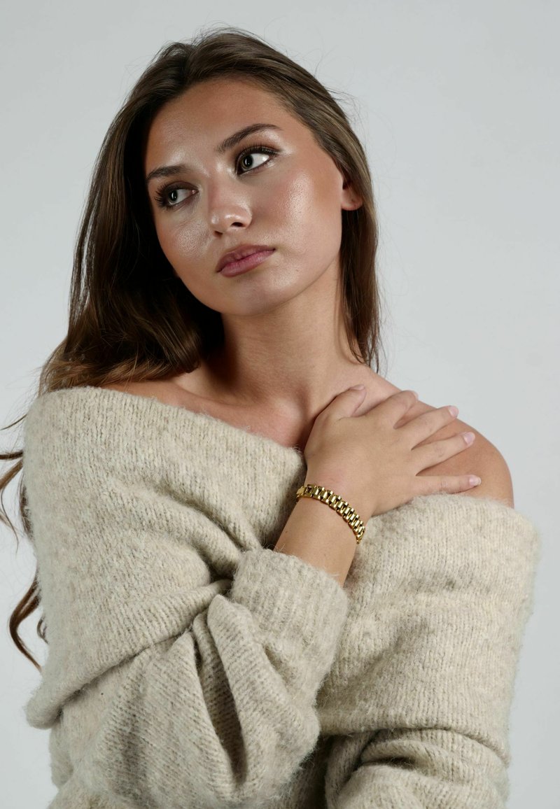 Gulda armband med sammanlänkade länkar, bärs på ett bar handled; modell i en mjuk beige, off-shoulder stickad tröja med en texturerad yta.