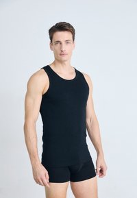 Homme aux cheveux bruns courts portant un débardeur noir et un short noir ajusté, debout contre un fond gris clair uni.