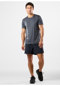 Harmaa urheilupaita, jossa on pystysuuntainen valkoinen logo, yhdistettynä mustiin shortseihin. Malli, joka pitää päällään mustia lenkkareita, seisoo yksinkertaista valkoista taustaa vasten.