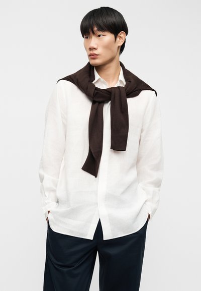 Chemise en lin blanche avec boutons sur le devant, manches longues, et un pull en tricot marron drapé sur les épaules. Un pantalon bleu marine foncé complète la tenue.