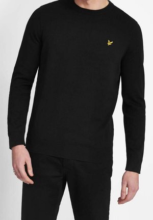 Uomo che indossa un maglione nero a maniche lunghe con un logo giallo di un uccello sul petto sinistro, abbinato a pantaloni neri.