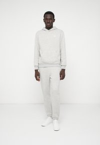 Pilkas džemperis su gobtuvu ir sportinės kelnės iš minkštos medžiagos, turinčios kengūros kišenę ir šonkaulių rankoves. Dėvima su baltais sportiniais bateliais.