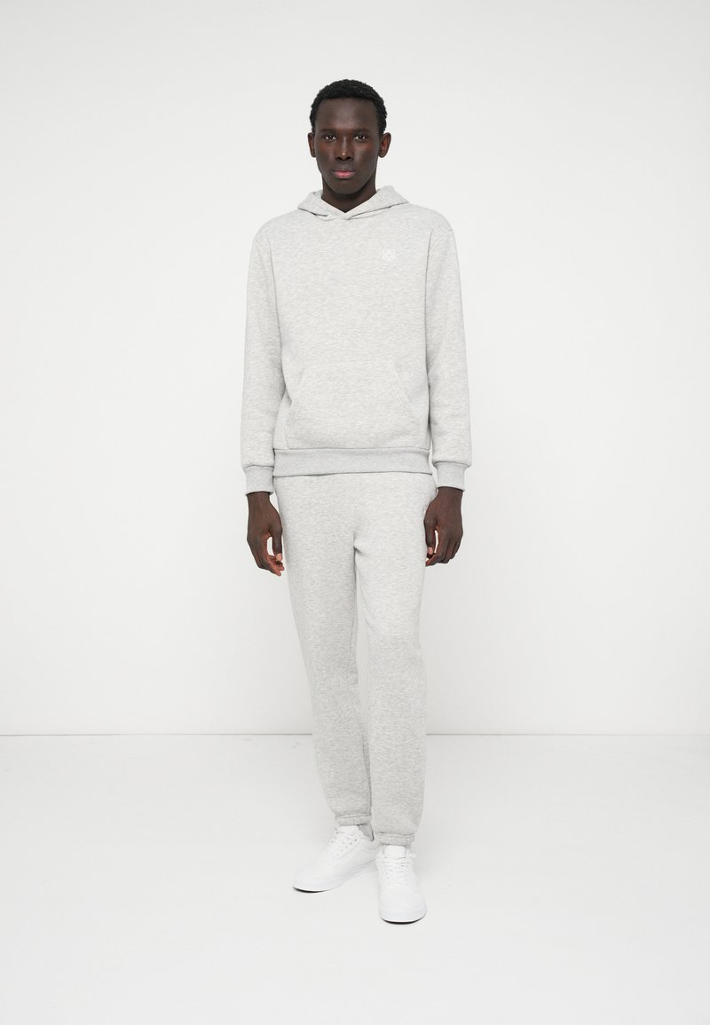 Pilkas džemperis su gobtuvu ir sportinės kelnės iš minkštos medžiagos, turinčios kengūros kišenę ir šonkaulių rankoves. Dėvima su baltais sportiniais bateliais.