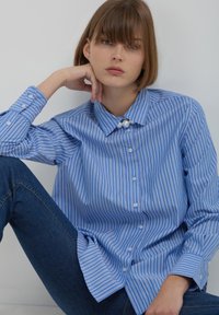 Young woman with short brown hair sitting against a white wall, wearing a blue and white striped shirt with pearl collar detail and dark jeans.