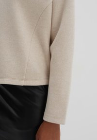 Maglione beige, con texture, a vestibilità ampia e maniche lunghe; presenta un orlo dritto e dettagli delle cuciture. Abbinato a una gonna nera e lucida.