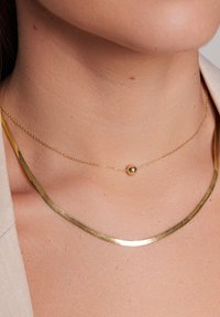 Naszyjnik w kolorze złotym z płaską, zakrzywioną belką oraz delikatnym łańcuszkiem z okrągłym akcentem. Gładka faktura i minimalistyczny design.