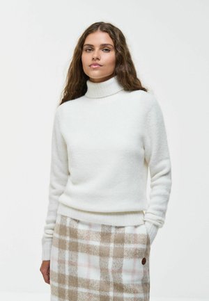 Une femme présente un pull à col roulé blanc et une jupe beige à carreaux sur un fond clair et uni.