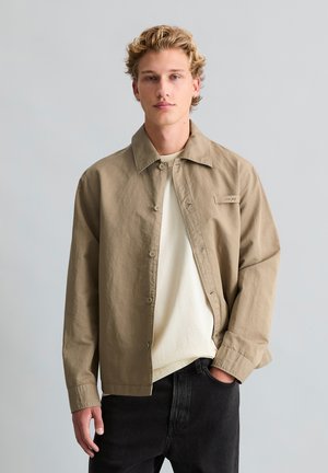 Młody mężczyzna z kręconymi blond włosami, ubrany w beżową koszulę zapinaną na guziki na kremową koszulkę i czarne dżinsy, stojący na jednolitym szarym tle.