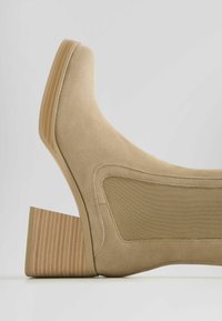 Béžová semišová kotníková obuv s obdélníkovým texturovaným podpatkem a elastickými bočními panely. Podpatky mají vroubkovaný design, který zvyšuje přilnavost a stabilitu.