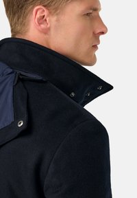 Vojaško sivi plašč iz volnenega mešanega materiala z visokim ovratnikom, ki ima satenasto ramenno ploščo in gumbe na zaklopu za prilagodljivost. Vidna mehka tekstura.