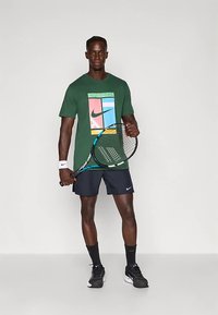 Man in green graphic t-shirt and black Nike shorts holding blue and black tennis racket with white wristband on white background.