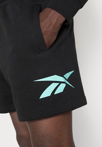 Czarne bawełniane szorty z boczną kieszenią i dużym turkusowym logo Reebok na dolnym brzegu. Tkanina ma gładką fakturę.