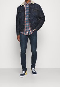 Uomo che indossa una giacca di jeans con colletto in shearling blu scuro, una camicia a quadri rossa e blu, jeans skinny neri e sneakers alto navy gialle.