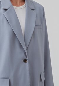 Blazer bleu clair en tissu lisse avec un bouton marron unique, deux poches avant et un col cranté.
