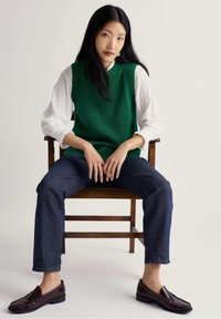 Chaleco de punto verde, blusa blanca con puños fruncidos, vaqueros azul oscuro y mocasines marrones. Sentada en una silla de madera sobre un fondo claro.