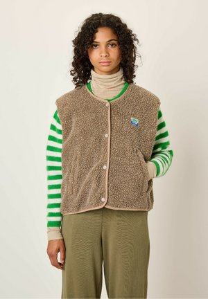 Brown fleece vest with beige trim and snap buttons, featuring side pockets and a colorful brand patch. Worn over a green striped sweater.