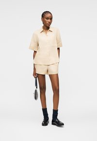 Femme aux cheveux tressés portant une chemise beige à manches courtes boutonnée et un short, des mocassins noirs avec des chaussettes foncées, tenant un petit sac à côté d'elle.
