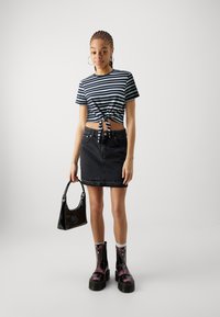 Svītrainā tumši zila krekla augšdaļa ar sasietu priekšpusi, melna denima mini svārki un spīdīga melna rokassoma. Modeli rotā melni un purpursarkani šņorētie zābaki.