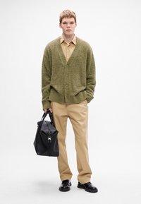 Olijfgroene fuzzy cardigan met knopen, beige overhemd met knopen, lichtbeige broek en een zwarte leren tas met een zilveren sluiting.