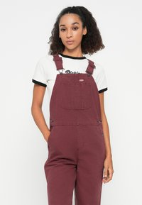 Salopette bordeaux avec bretelles ajustables, poche frontale et boutons sur les côtés. Associée à un t-shirt blanc avec col en rond noir et texte.