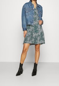 Giacca di jeans con chiusura a bottoni, due tasche sul petto; vestito verde floreale con orlo svasato; stivali neri alla caviglia con punta appuntita.