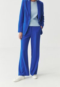 Královská modrá na míru ušitá bunda a odpovídající široké kalhoty, doplněné světle modrým topem a bílými teniskami. Hladká tkanina, čisté linie, formální design.