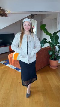 Νεαρή γυναίκα με μακριά μαλλιά, φορώντας λευκό σακάκι, μαύρο φόρεμα, γκρι παπούτσια και λευκή κορδέλα, στέκεται σε ένα φωτεινά φωτισμένο σαλόνι.