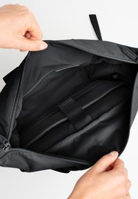 Interno di una borsa nera con zip, dotato di scomparti imbottiti e cintura elastica, realizzata in plastica riciclata. Texture liscia, senza motivi visibili.