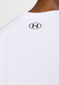 Baltas medvilninis marškinėlis suapvalintu kaklu, ant nugaros yra juodas Under Armour logotipas. Audinys atrodo lygus ir lengvas.