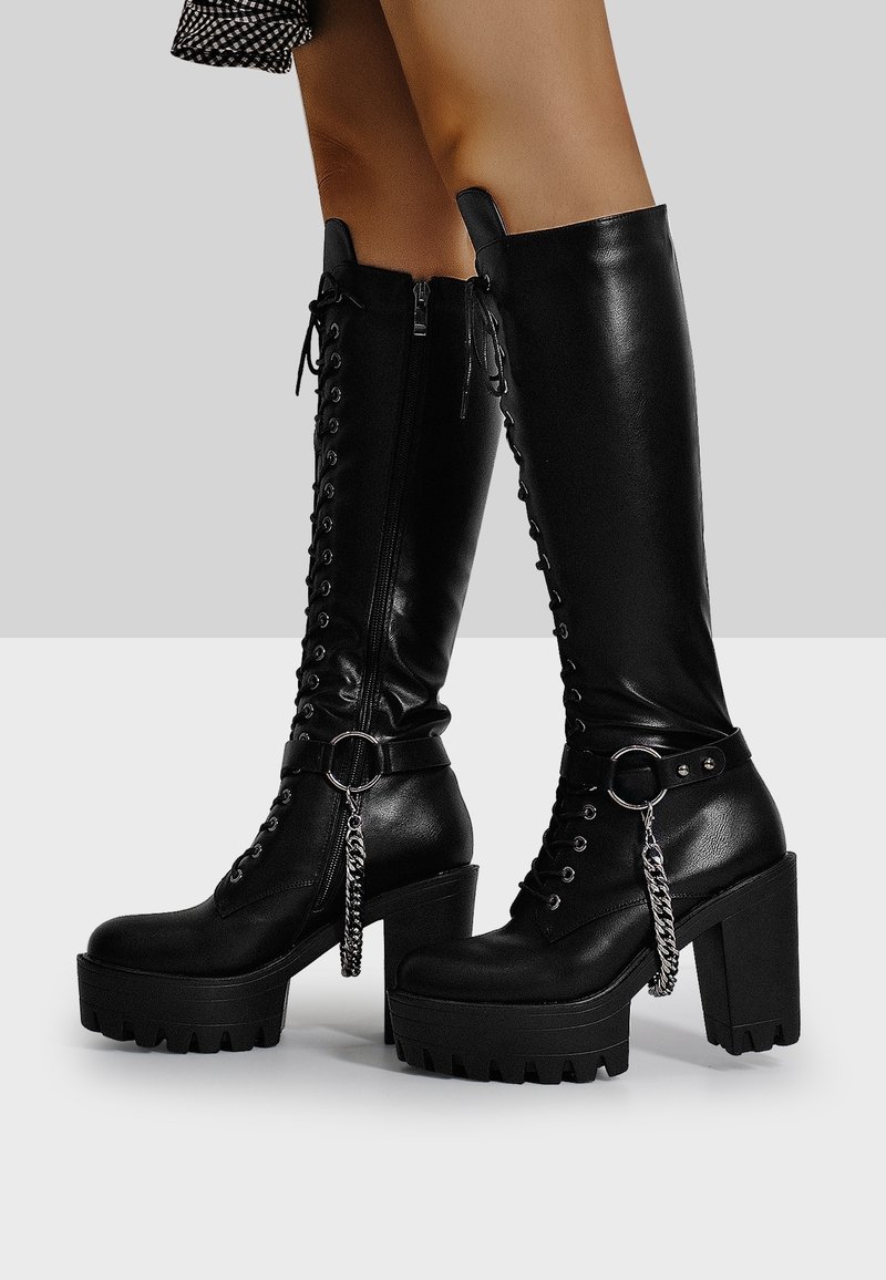 Black knee-high boots with chunky heels, lace-up front, metal ring accents, and a side zipper, featuring a textured finish and chain detail.