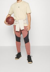 Camiseta beis para hombres con un logo negro, combinada con pantalones de chándal en bloques de color rojo y negro. Sosteniendo un baloncesto, con zapatillas negras.