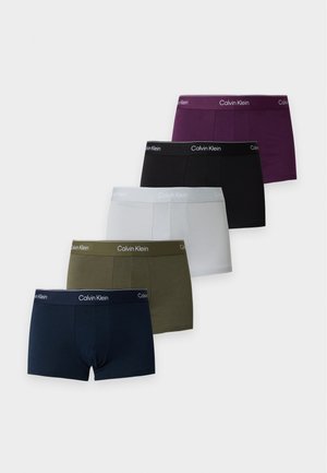 Homme musclé debout pieds nus portant un boxer noir Calvin Klein, bras croisés, sur un fond blanc uni.