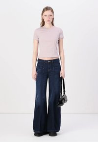 T-shirt fitted di un rosa chiaro, jeans a vita alta blu scuro a zampa d'elefante, piccole tasche frontali, borsa nera, scarpe nere basse, sfondo neutro.